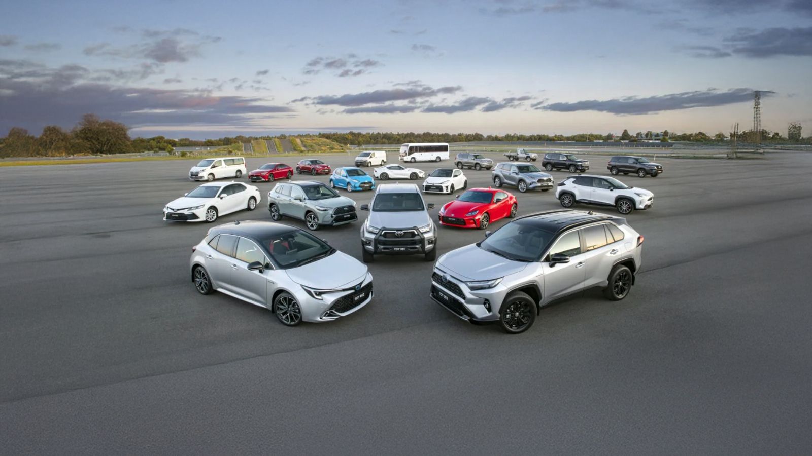 Több különböző Toyota személyautó és SUV modell parkol egy kör alakú elrendezésben egy nyitott tesztpályán, naplemente idején.
