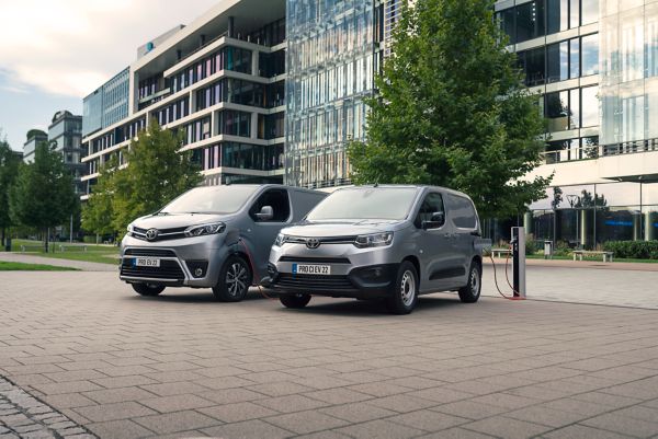 Ein Proace City Electric und Proace City Verso Electric nebeneinander an der Ladestation geparkt vor einer modernen Häuserfront.