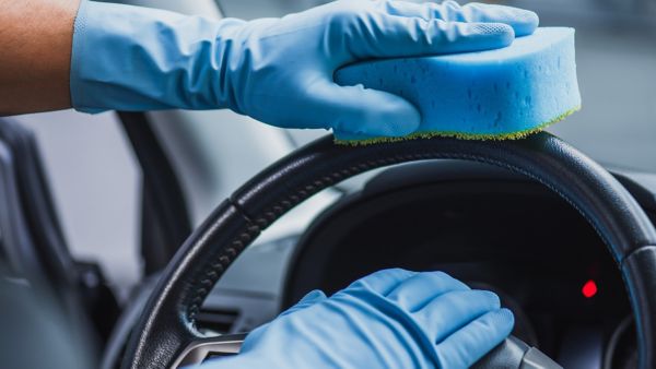 Die Nahaufnahme zeigt, wie ein Lenkrad mit einem blauen Schwamm und Handschuhen gründlich gereinigt wird.