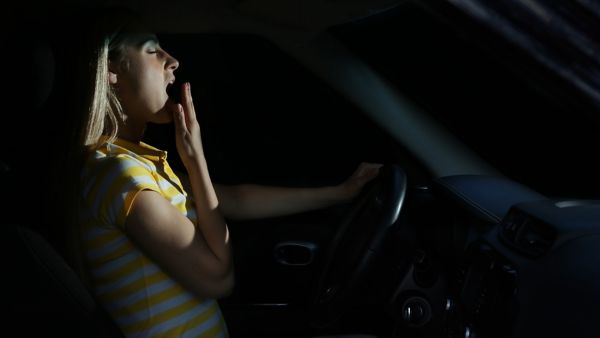 Im Scheinwerferlicht ist eine junge Autofahrerin zu sehen, die sich mit geschlossenen Augen die Hand vor den Mund hält, während sie gähnt.