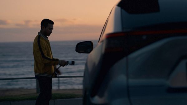 Mann in gelber Jacke steht bei Sonnenuntergang neben einem Auto am Meer