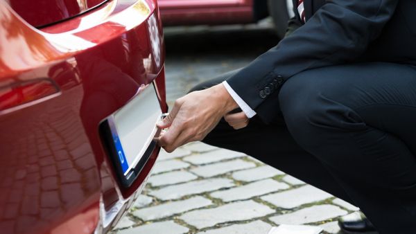 Ein Mann entfernt auf einem Parkplatz das hintere Nummernschild eines Fahrzeugs, was auf eine bevorstehende Kfz-Abmeldung hinweist.