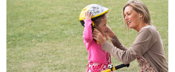 Eine Frau hilft einem jungen Mädchen, ihren gelben Helm zu richten, während sie sich auf einer Wiese auf das Spielen im Freien mit einem Roller vorbereitet.