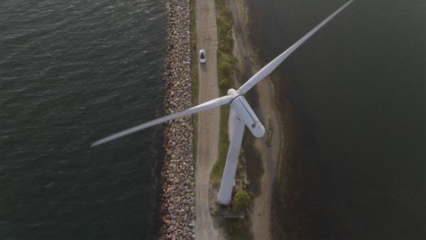 Luftaufnahme einer Windkraftanlage an der Küste entlang einer Straße, mit einem Auto auf dem Weg und Wasser auf einer Seite.