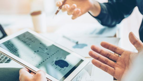 Detailansicht auf zwei Hände, die ein Tablet auf dem Graphen dargestellt sind und einen Stift halten. Die zwei Personen scheinen in einem Meeting zu sein. 
