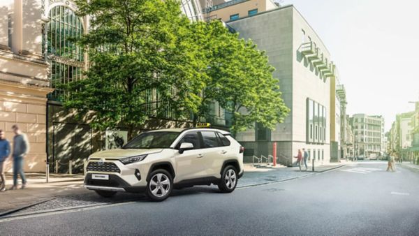 Ein weißer SUV, der auf einer Straße in der Stadt geparkt ist, umgeben von grünen Bäumen und modernen Gebäuden, mit Fußgängern, die im Hintergrund vorbeilaufen.