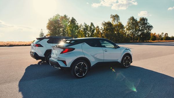 Ein Toyota C-HR Hybrid und RAV4 Hybrid nebeneinander, die auf einer gepflasterten Fläche geparkt sind unter einem klaren blauen Himmel, umgeben von Bäumen.