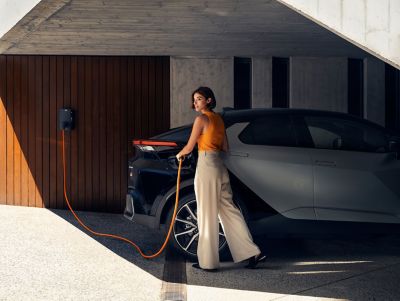 Frau lädt ihren Toyota C-HR über ein orangenes Kabel an einer Wallbox in einer überdachten Einfahrt auf.