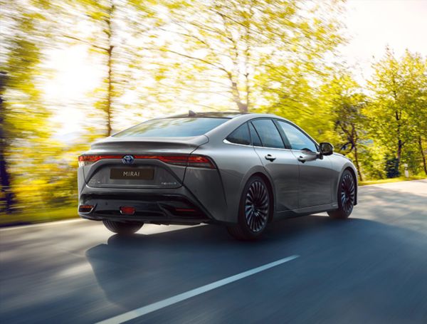 Ein grauer Toyota Mirai fährt auf einer von Bäumen gesäumten Straße. Sonnenlicht scheint durch die Bäume im Hintergrund.