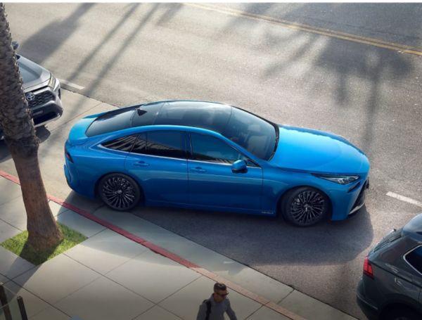 Ein blauer Mirai parkt seitlich zwischen zwei weiteren Autos an eine Straße ein. Daneben ist ein Baum und eine Person angeschnitten zu sehen.