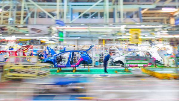 Autokarosserien bei der Fertigung in einer Halle mit einzelnen Mitarbeitern, bis auf eine blaue Karosserie im Zentrum ist alles unscharf