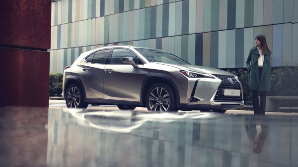 Eine elegante Frau in einem grünen Mantel steht neben einem silbernen Lexus UX, der vor einem bunten Gebäude geparkt ist. Im Wasser sind Spiegelungen des Autos und der Umgebung zu sehen.