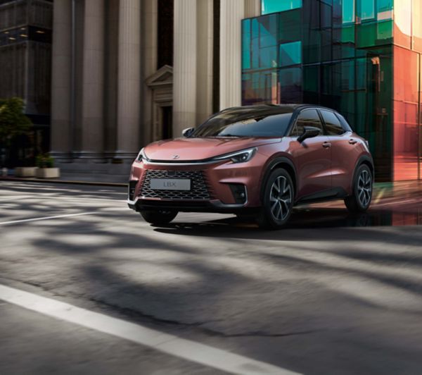 Ein Lexus LBX wird in Bewegung auf einer städtischen Straße gezeigt, mit Glasgebäuden und Grünflächen im Hintergrund