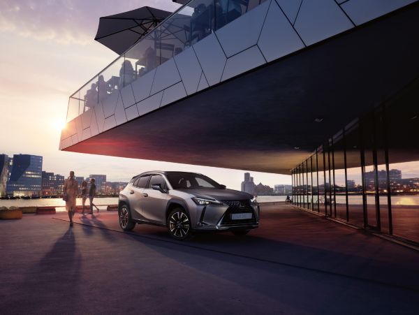 Ein silberner Lexus UX ist bei Sonnenuntergang in einer städtischen Umgebung geparkt, in der Menschen spazieren gehen und moderne Gebäude am Wasser stehen.