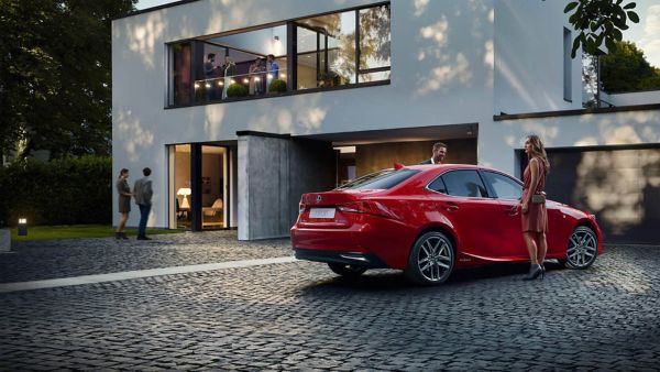 Ein Paar steht in der Nähe eines roten Lexus in einer Einfahrt, während sich Freunde auf einem Balkon in einer modernen Wohnumgebung treffen.