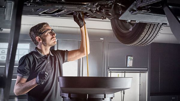 Ein Lexus-Mechaniker mit Handschuhen und Schutzbrille prüft in einer Werkstatt das Öl, das von der Unterseite eines Autos in eine schwarze Auffangwanne abläuft
