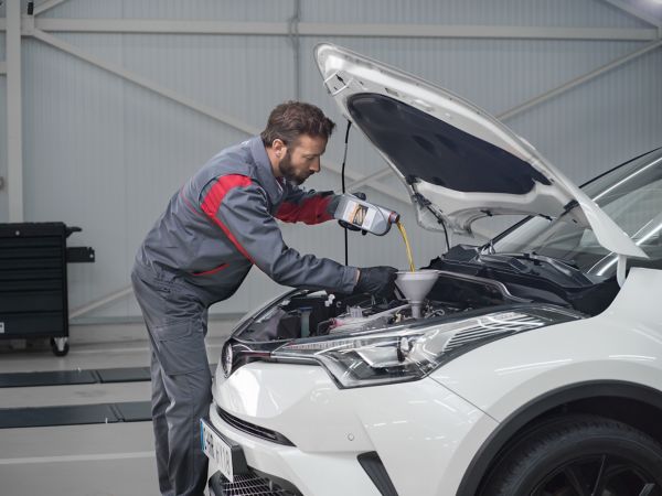 Ein Toyota-Werkstattmitarbeiter in grau-roter Arbeitskleidung gießt Öl in den Motor eines weißen Toyotafahrzeugs.