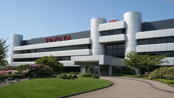 Modernes Toyota-Gebäude mit einem einladenden Eingang, umgeben von grünem Gras, Bäumen und blühenden Kirschbäumen unter einem strahlend blauen Himmel.