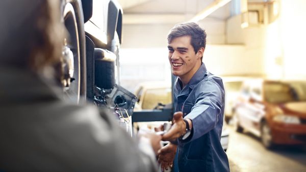 Ein lächelnder junger Mechaniker streckt seine Hand für ein Werkzeug aus, während er neben einem Fahrzeug in einer Werkstatt steht