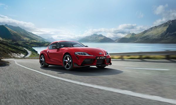 Ein roter GR Supra fährt auf einer Landstraße vor einem Bergsee. In Hintergrund sind ein Gebirge zu sehen, dass von Wolken bedeckt wird.