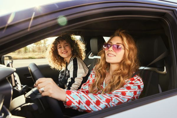 Zwei lächelnde Frauen in einem Auto, eine von ihnen fährt mit Sonnenbrille und genießt einen sonnigen Tag auf der Straße. Die Atmosphäre ist freundlich und unbeschwert.