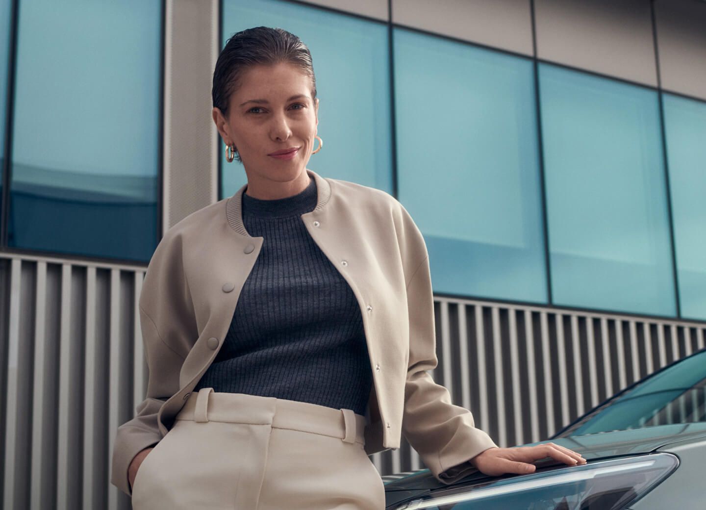 Eine Frau lehnt an einer Motorhaube eines Autos, während im Hintergrund ein modernes Gebäude steht