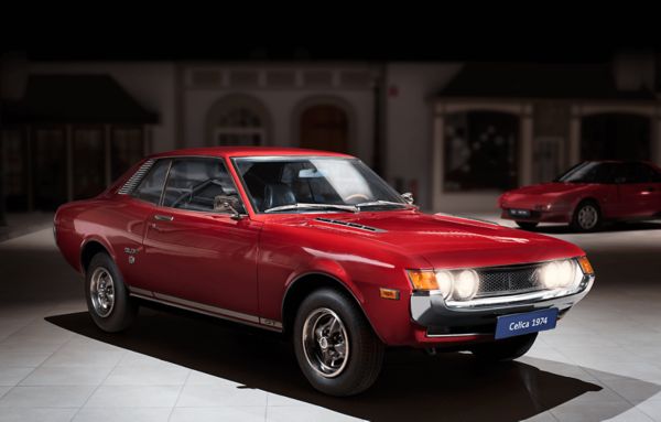 Roter Toyota Celica von 1974 mit leuchtenden Scheinwerfern, von schräg vorn im Spot-Licht in einer abgedunkelten Ausstellungshalle 