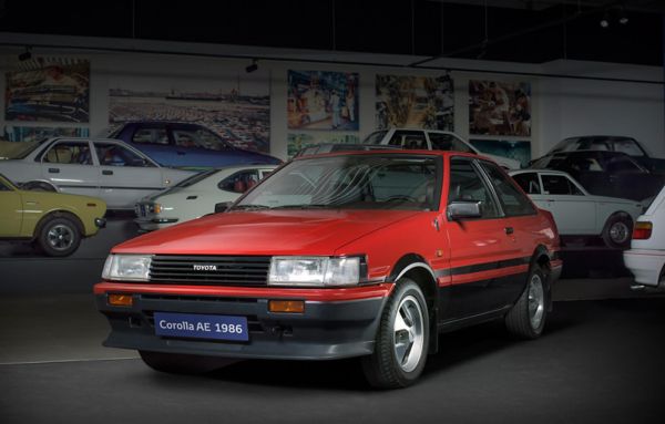 Ein roter Toyota Corolla AE aus dem Jahr 1986 schräg von vorn in einer Ausstellungshalle, im Hintergrund weitere Modelle