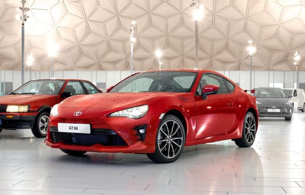 Ein roter Toyota GT86 mit leuchtenden Scheinwerfern von schräg vorne in einer hell ausgeleuchteten Ausstellungshalle, im Hintergrund weitere Modelle