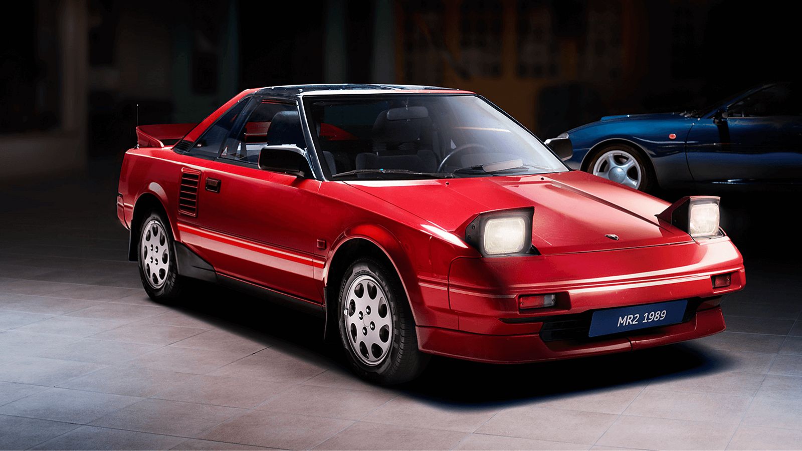 Roter Toyota MR2 von 1989 mit aufgeklappten Scheinwerfern, von schräg vorn im Spot-Licht in einer abgedunkelten Ausstellungshalle 