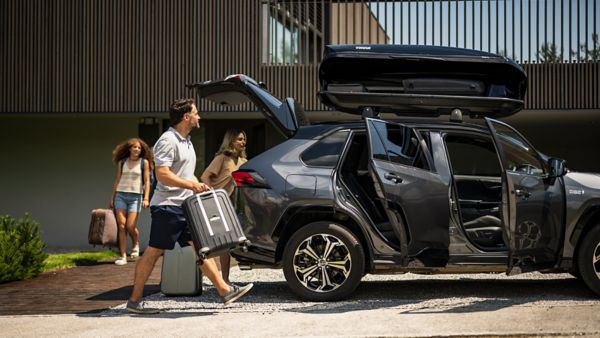 Grauer RAV4 mit Gepackträger und offenen Türen steht vor einem Haus. Eine Familie räumt gerade Gepäck hinein.