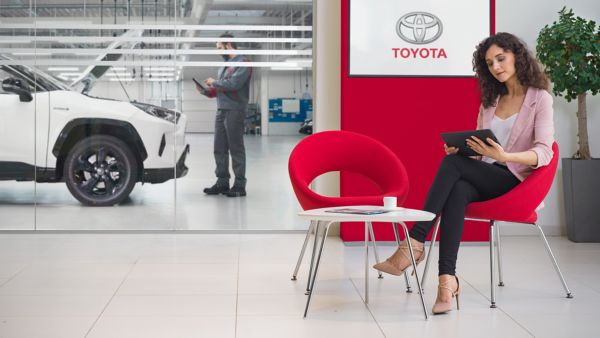 Eine Frau sitzt mit einem Tablet in einer Toyota Fachwerkstatt mit einem weißen Auto im Hintergrund und einem Mechaniker, der das Fahrzeug inspiziert.