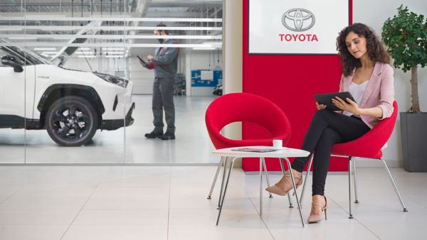 Eine Frau, die in einem roten Stuhl sitzt und ein Tablet in einem Toyota-Autohaus benutzt, mit einem weißen Fahrzeug und einem Mechaniker im Hintergrund.