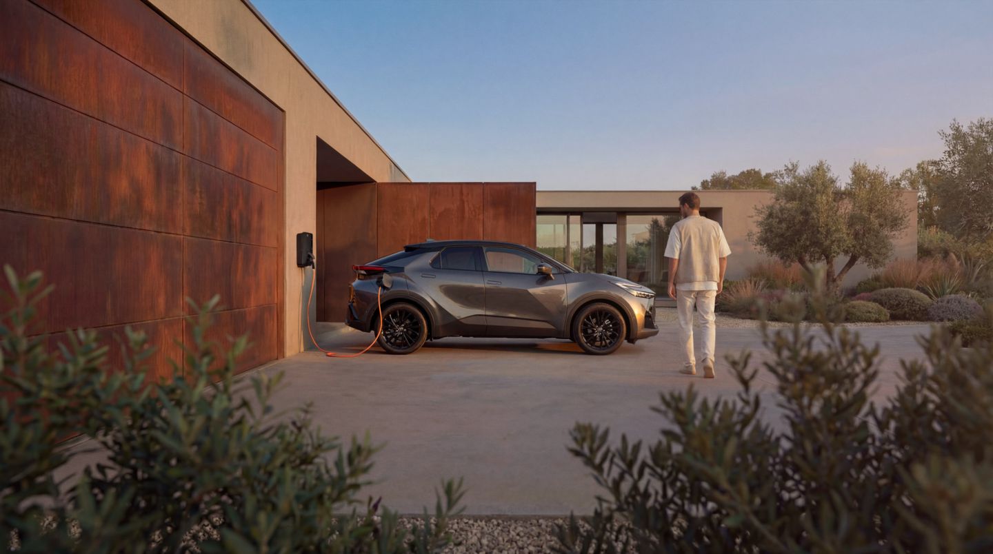 Ein grauer Toyota C-HR steht vor einem modernen Haus und lädt an einer Wallbox; eine Person geht über die Einfahrt auf das Fahrzeug zu