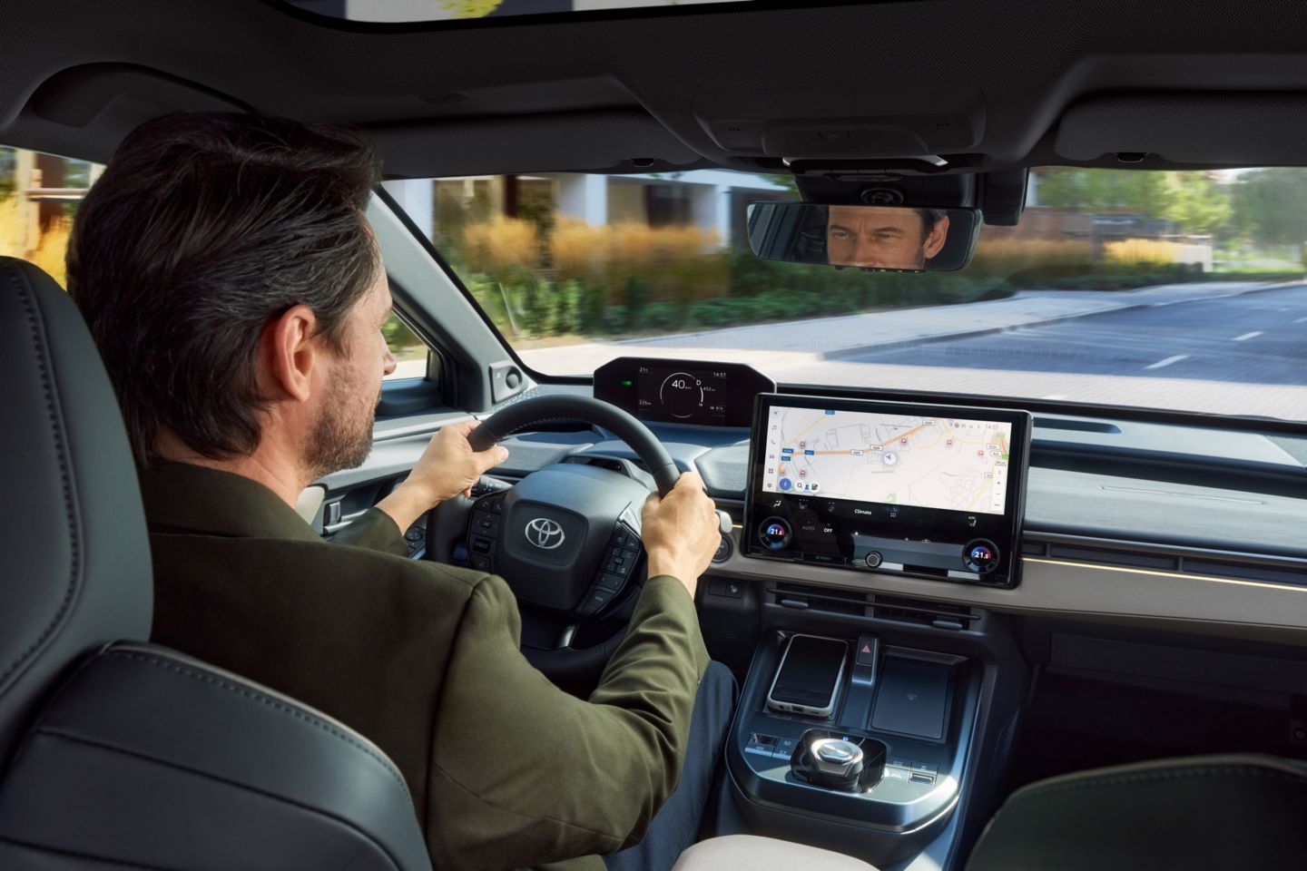Blick von der Rückbank auf den Fahrersitz: Eine Person steuert das Auto, vorne ist ein Navigationsbildschirm zu sehen, draußen verläuft eine ruhige Straße durch ein Wohngebiet.