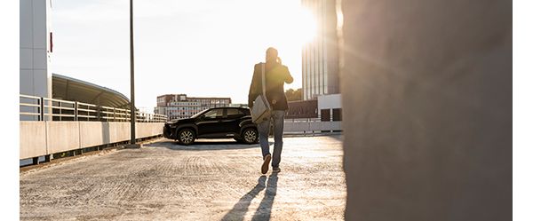 Auf einem Parkdeck geht eine Person mit Smartphone am Ohr und weißer Tasche der Sonne entgegen zu seinem geparkten Toyota.