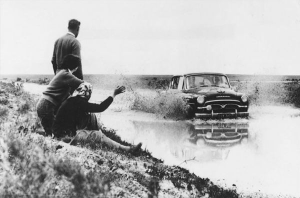 Schwarz-weiß Bild eines Oldtimers, der durch eine Pfütze auf einer Landstraße fährt, während zwei Personen von der Seite zuschauen.