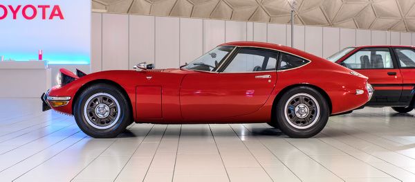 Seitenansicht eines roten Toyota 2000GT von 1968 mit sportlicher Silhouette und aufgeklappten Scheinwerfern in einer hellen Ausstellungshalle