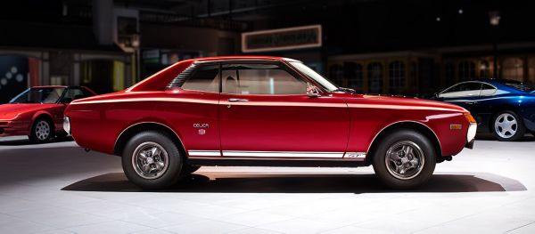 Seitenansicht eines roten Toyota Celica von 1974 im Spot-Licht in einer abgedunkelten Ausstellungshalle, im Hintergrund weitere Fahrzeuge