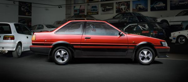 Seitenansicht eines roten Toyota Corolla AE aus dem Jahr 1986 im Spot-Licht in einer abgedunkelten Ausstellungshalle, im Hintergrund weitere Fahrzeuge