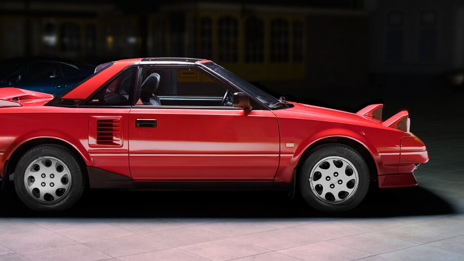 Seitenansicht eines roten Toyota MR2 mit hochgeklappten Scheinwerfern im Spot-Licht in einer abgedunkelten Ausstellungshalle