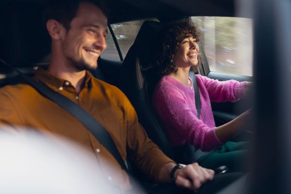 Eine Frau am Steuer eines fahrenden Autos, ein Mann als Beifahrer, beide blicken lachend nach vorn