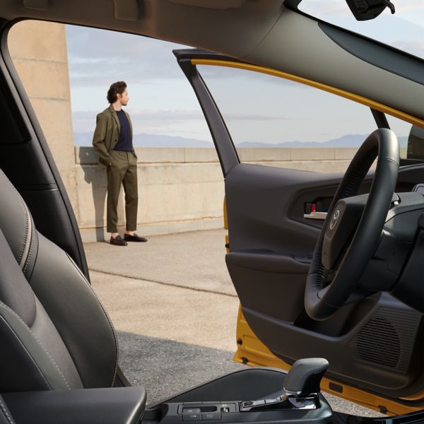 Blick auf den leeren Fahrersitz und die geöffnete Fahrertür eines Toyota, im Hintergrund schaut ein junger Mann auf eine Bergkette in der Ferne