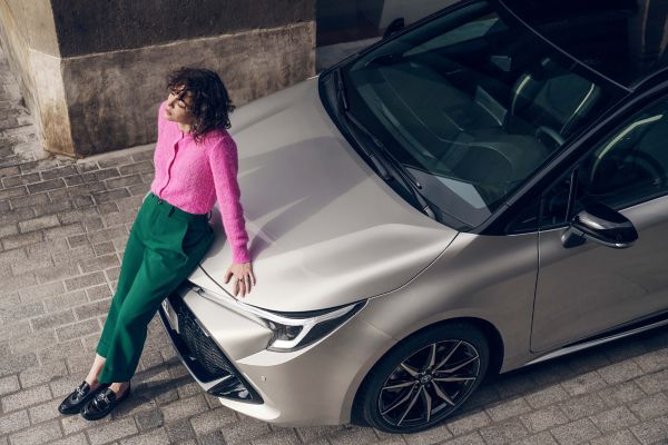 Eine junge Frau lehnt an der Motorhaube eines silbernen Corolla und genießt mit geschlossenen Augen den Sonnenschein.