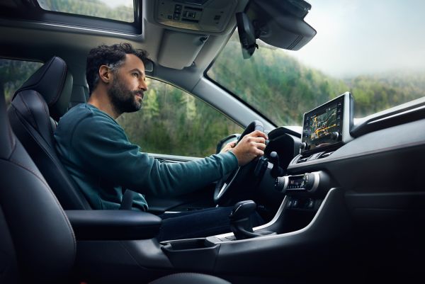 Ein Mann fährt ein modernes Auto und konzentriert sich auf die Straße vor ihm. Das Auto ist mit einem digitalen Display und einem Panoramadach ausgestattet, durch das man grüne Hügel sehen kann.