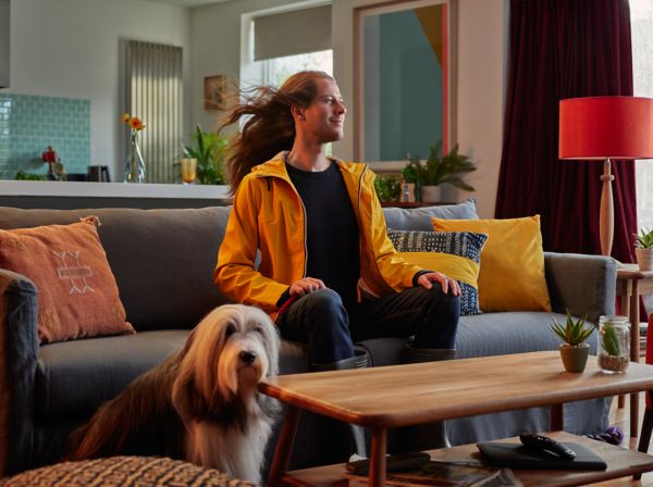 Eine Person in einer gelben Jacke sitzt auf einem Sofa in einem gemütlichen Wohnzimmer, neben dem Couchtisch steht ein Hund.