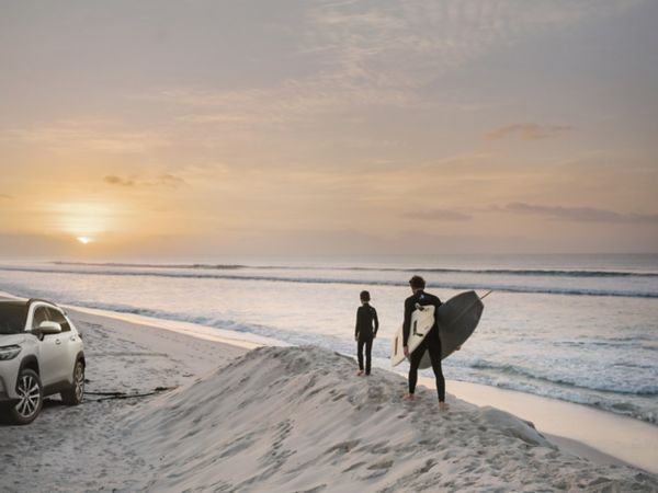 zwei Surfer laufen während des Sonnenuntergangs am Strand entlang, während links von ihnen in weißer SUV von Toyota parkt.