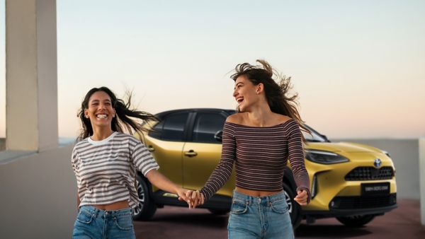 Zwei Frauen, die freudig Hand in Hand gehen, mit einem gelben Auto im Hintergrund. Die Szene fängt einen lebendigen, sorgenfreien Moment ein.