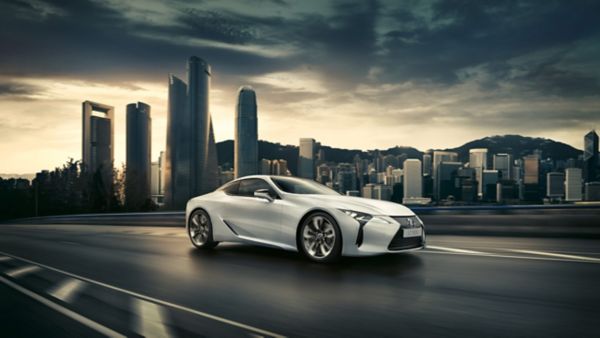 Ein eleganter weißer Lexus LC fährt auf einer Straße mit einer modernen Skyline im Hintergrund unter einem dramatischen Himmel, der eine Mischung aus Luxus und Stadtlandschaft darstellt.