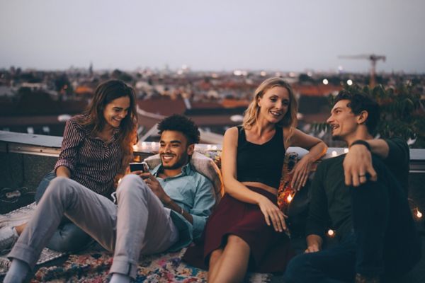 Eine Gruppe von vier Freunden verbringt einen lebhaften Abend auf einem Dach und lacht und plaudert mit der mit funkelnden Lichtern geschmückten Skyline der Stadt im Hintergrund.